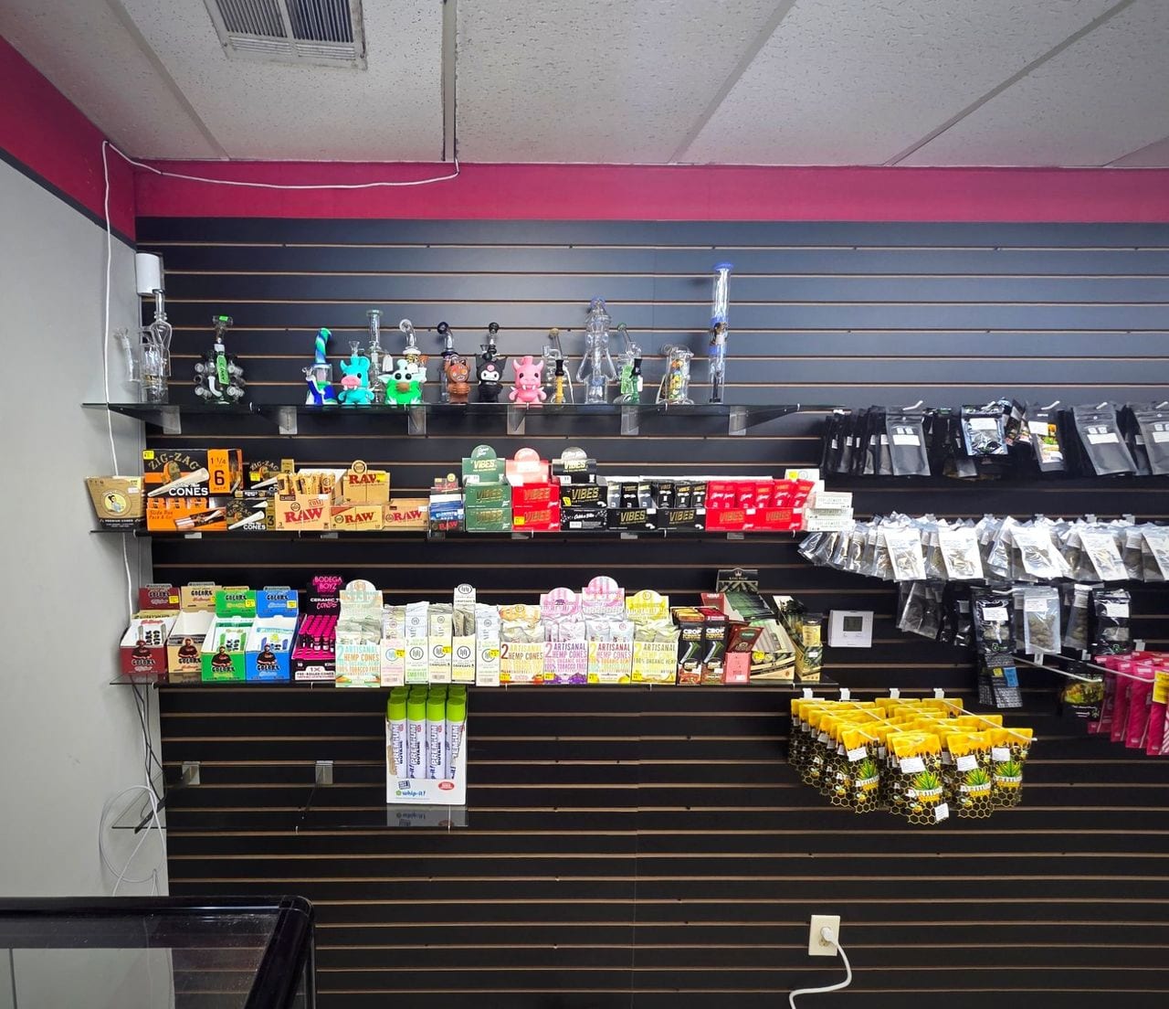 A glass shelf of pipes and water pieces above a wall of rolling papers and cones.